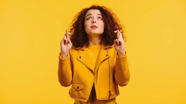Joven mujer rizada con los dedos cruzados pidiendo deseo y mirando hacia arriba aislado en amarillo - foto de stock