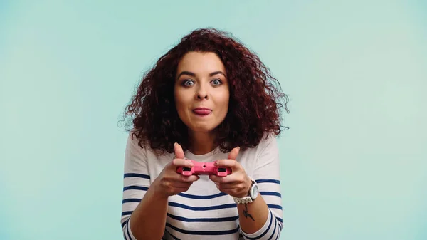 KYIV, UKRAINE - JUNE 30, 2021: young curly woman holding pink joystick and sticking out tongue while playing video game isolated on blue — Stock Photo