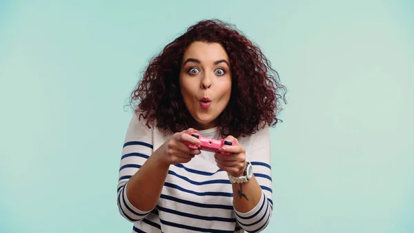 QUIIV, UCRÂNIA - JUNHO 30, 2021: jovem mulher surpresa segurando joystick rosa e jogando videogame isolado em azul — Fotografia de Stock