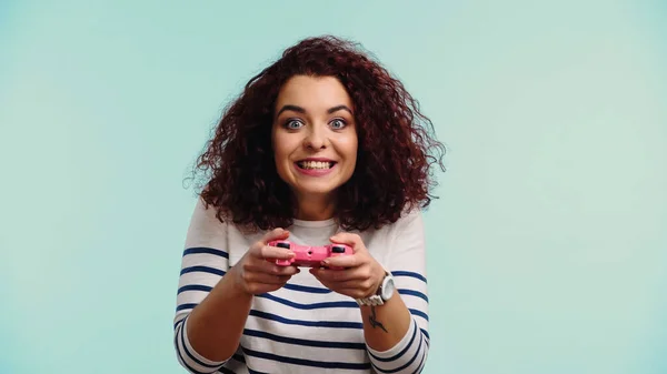 KYIV, UCRANIA - 30 de junio de 2021: una joven feliz sosteniendo un joystick rosa y jugando un videojuego aislado en azul - foto de stock