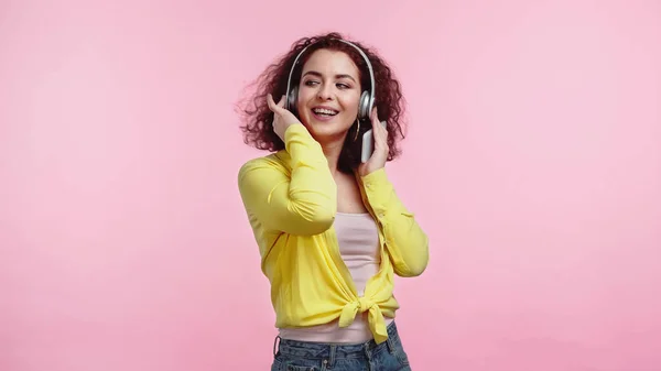 Joven mujer alegre en auriculares inalámbricos sosteniendo teléfono inteligente aislado en rosa - foto de stock