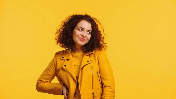 Усміхнена молода жінка в шкіряній куртці стоїть з рукою на стегні ізольовано на жовтому — стокове фото