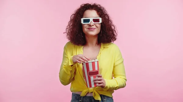 Feliz mujer joven en gafas 3d sosteniendo palomitas de maíz cubo aislado en rosa - foto de stock