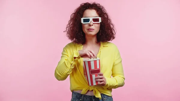 Curly young woman in 3d glasses holding popcorn bucket isolated on pink — Stock Photo