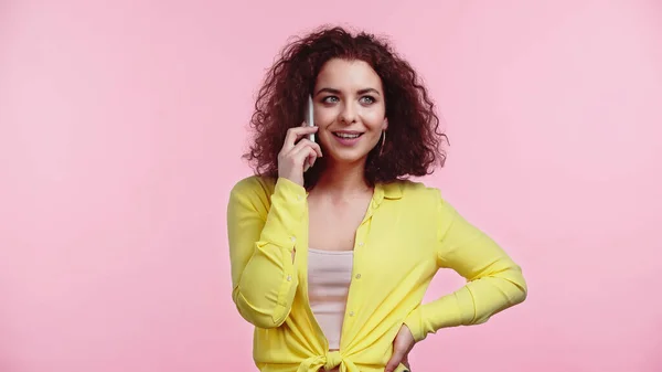 Cheerful woman standing with hand on hip while talking on cellphone isolated on pink — Stock Photo