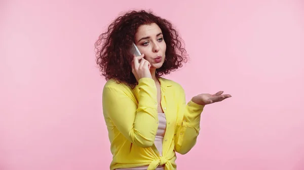 Joven mujer rizada gesto mientras habla en el teléfono celular aislado en rosa - foto de stock