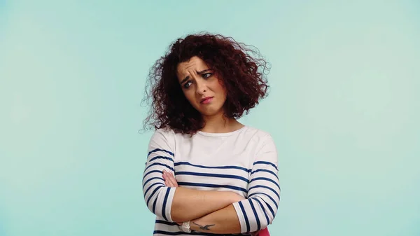 Sad young woman in striped long sleeve shirt standing with crossed arms isolated on blue — Stock Photo