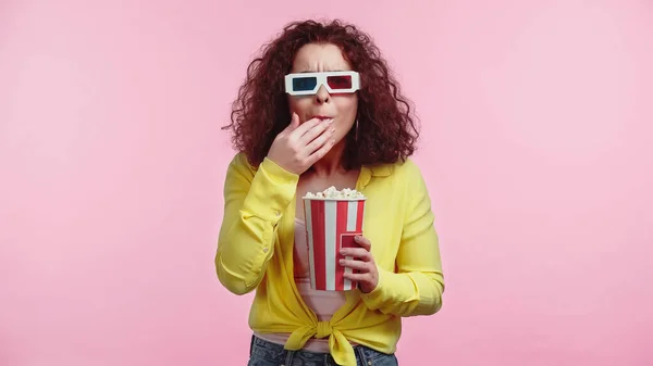 Mujer rizada en gafas 3d sosteniendo cubo mientras come palomitas de maíz aislado en rosa - foto de stock