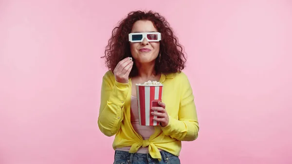 Feliz joven en gafas 3d sosteniendo cubo mientras come palomitas de maíz aislado en rosa - foto de stock