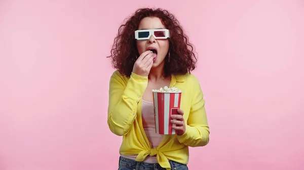 Curly young woman in 3d glasses with open mouth eating popcorn isolated on pink — Stock Photo