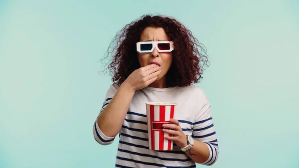 Curly young woman in 3d glasses eating popcorn isolated on blue — Stock Photo