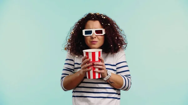 Curly young woman in 3d glasses with bucket and popcorn in hair isolated on blue — Stock Photo