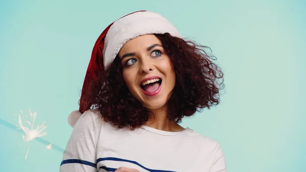 Stupito giovane donna in cappello di Babbo Natale vicino sparkler isolato su blu — Foto stock