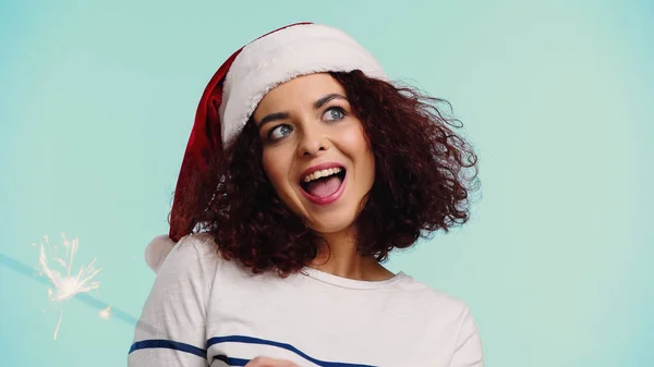 Amazed young woman in santa hat near sparkler isolated on blue — Stock Photo