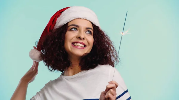 Mulher positiva em santa chapéu segurando sparkler isolado em azul — Fotografia de Stock