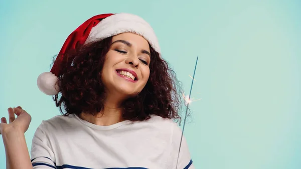 Curly and positive woman in santa hat holding sparkler isolated on blue — Stock Photo