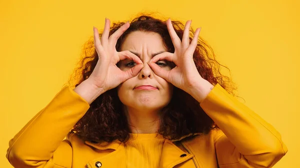 Young curly woman imitating eyeglasses with hands and grimacing isolated on yellow — Stock Photo