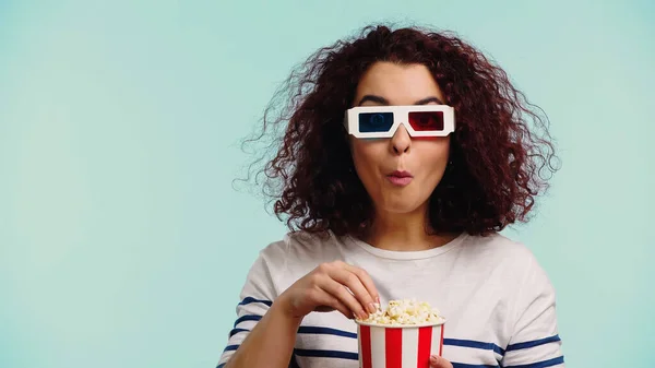 Curly woman in 3d glasses eating popcorn while holding bucket isolated on blue — Stock Photo