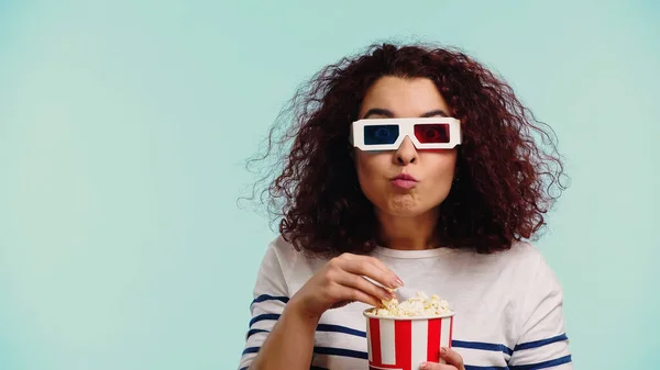 Mujer joven rizado en gafas 3d comer palomitas de maíz mientras sostiene cubo aislado en azul - foto de stock