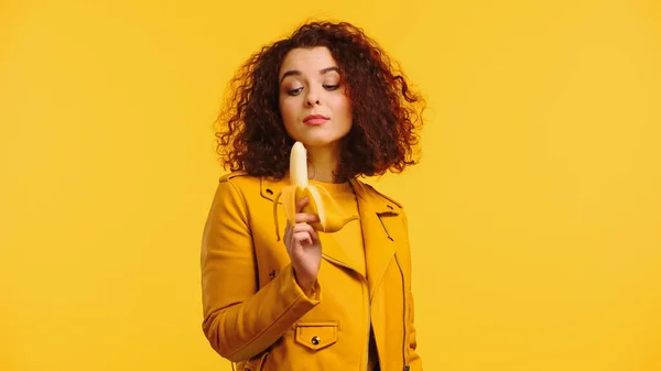 Giovane donna riccia in giacca di pelle guardando banana isolata su giallo — Foto stock