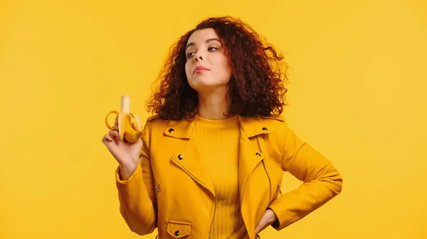 Young curly woman in leather jacket holding ripe banana while standing with hand on hip isolated on yellow — Stock Photo