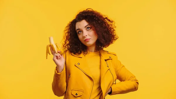 Femme bouclée en veste tenant banane mûre tout en se tenant avec la main sur la hanche isolé sur jaune — Photo de stock