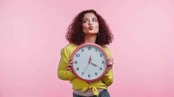 Кучерява жінка тримає годинник і кидає губи ізольовані на рожевому — стокове фото
