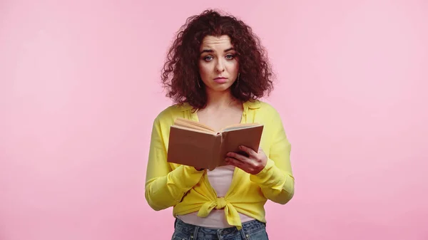 Lockige und verärgerte Studentin hält Buch isoliert auf rosa — Stockfoto