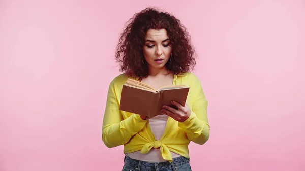 Rizado y sorprendido estudiante lectura libro aislado en rosa - foto de stock