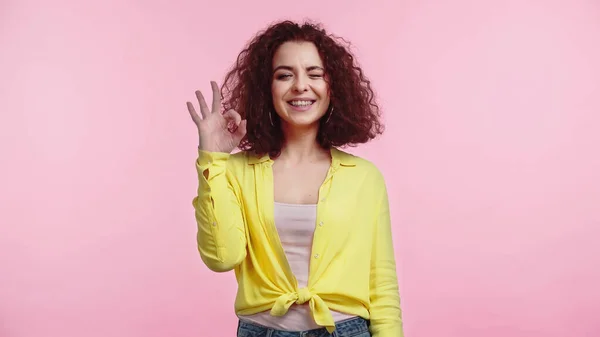 Alegre joven y rizado mujer mostrando ok signo mientras guiño aislado en rosa - foto de stock