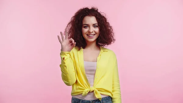 Pleased young and curly woman showing ok sign isolated on pink — Stock Photo