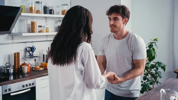 Sorridente homem em t-shirt segurando mão de morena namorada na cozinha — Fotografia de Stock