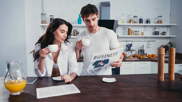 Jovens amantes com xícaras de café lendo jornal de negócios na cozinha — Fotografia de Stock