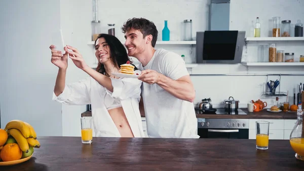 Sexy mujer en blanco camisa y sujetador tomando selfie con novio celebración de panqueques - foto de stock