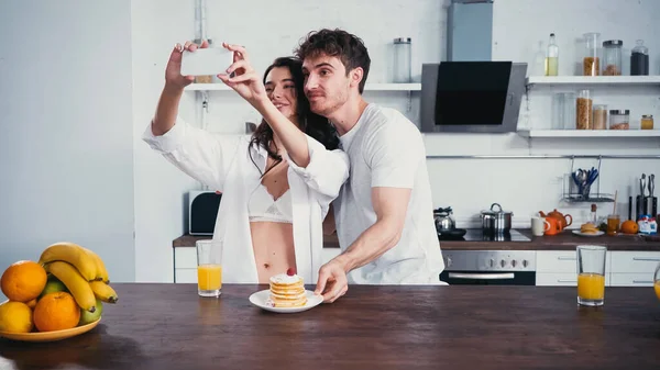 Lächelnde Frau in Hemd und BH macht Selfie mit Freund in der Nähe von Pfannkuchen — Stockfoto