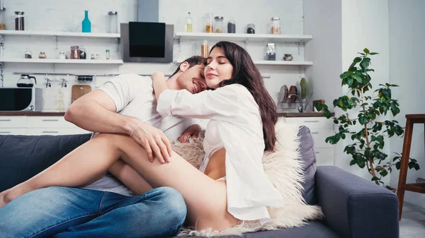 Smiling man in jeans embracing hot woman with naked legs on sofa in kitchen — Stock Photo