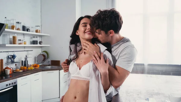 Joven abrazando sexy, mujer sonriente en camisa y sujetador en la cocina - foto de stock