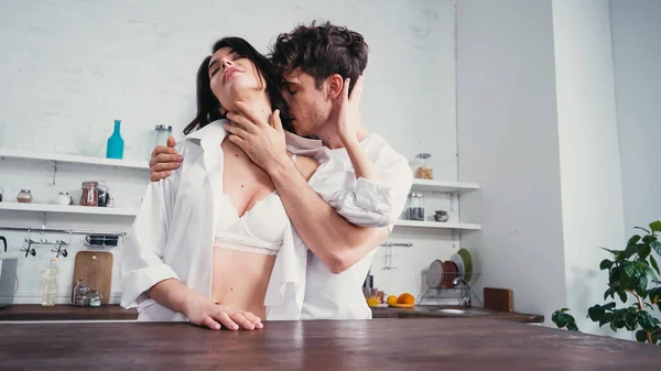 Passionate woman in unbuttoned shirt and bra near man embracing and kissing her in kitchen — Stock Photo