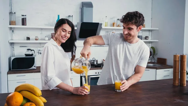 Homem feliz derramando suco de laranja fresco perto de mulher morena na camisa branca — Fotografia de Stock