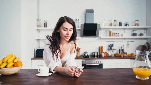 Mulher sedutora em camisa branca e mensagens de sutiã no smartphone perto de café e frutas — Fotografia de Stock