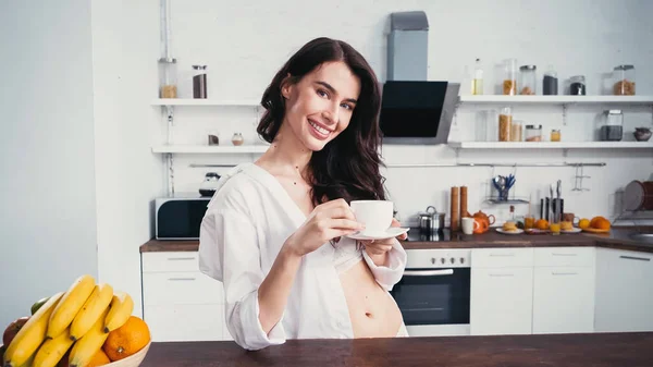 Сексуальная женщина с чашкой кофе улыбаясь в камеру возле свежих фруктов на кухне — стоковое фото