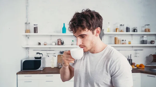 Young man in white t-shirt drinking morning coffee in kitchen — Stock Photo