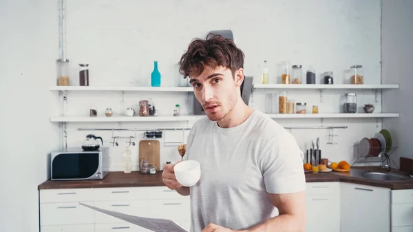 Homem com jornal e xícara de café olhando para a câmera na cozinha — Fotografia de Stock