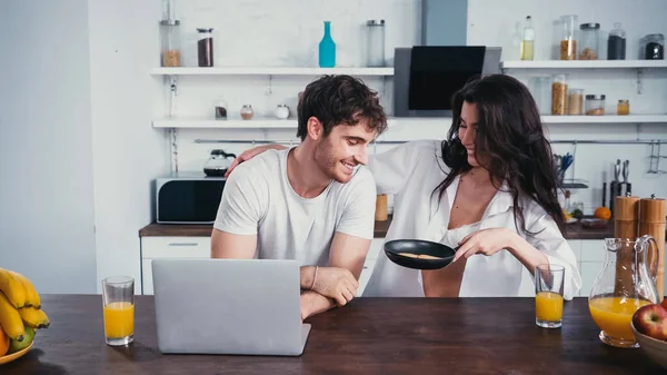 Sexy Frau im aufgeknöpften Hemd zeigt Mann in der Küche Bratpfanne neben Laptop — Stockfoto