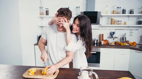 Cheerful woman in white shirt covering eyes of man cutting orange in kitchen — Stock Photo