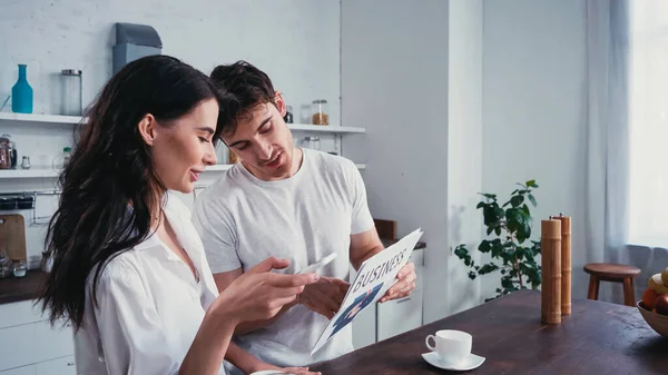 Мужчина показывает деловую газету женщине в расстегнутой рубашке, используя смартфон на кухне — стоковое фото