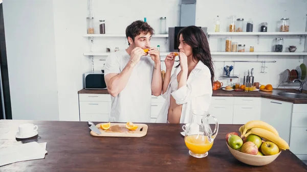 Hombre joven y mujer sexy en sujetador y camisa comiendo naranja madura en la cocina - foto de stock