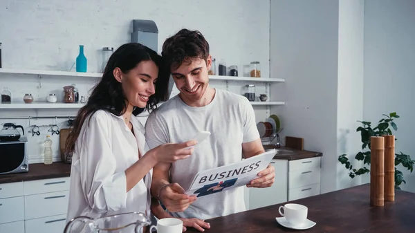 Smiling man showing business newspaper to pretty girlfriend using smartphone in kitchen — Stock Photo