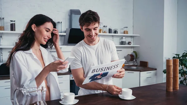 Smiling couple using smartphone and reading business newspaper in kitchen — Stock Photo