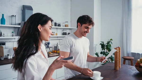 Bonita mujer sosteniendo teléfono inteligente cerca sonriente hombre leyendo periódico de la mañana - foto de stock